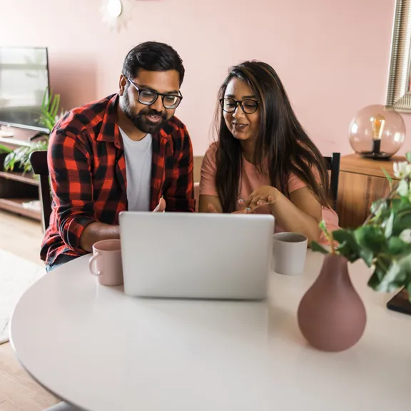 Smiling couple seated at a round table looking at a laptop with mugs beside them