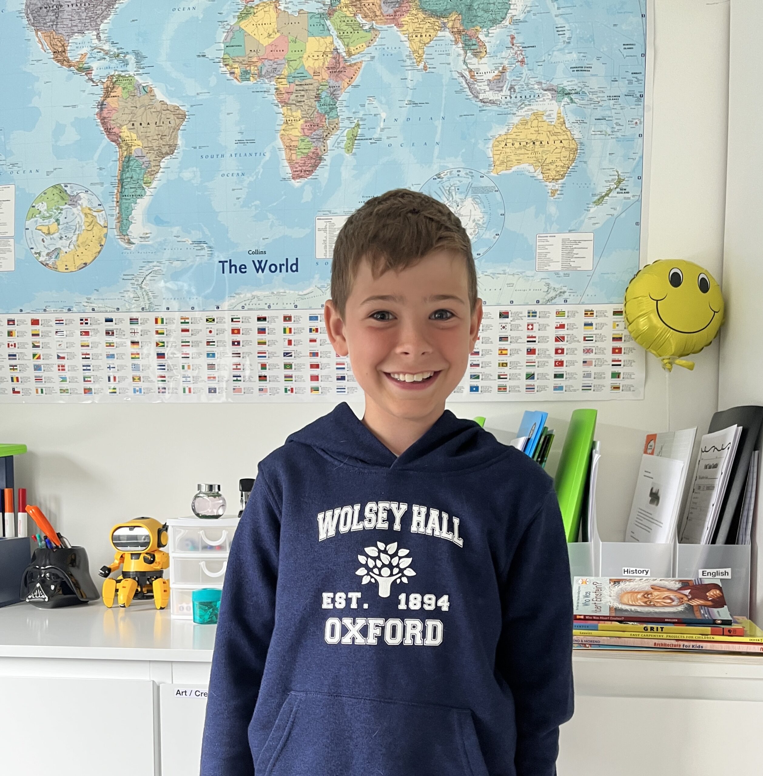 Happy student wearing a Wolsey Hall Oxford hoodie, standing in front of a colourful world map at home