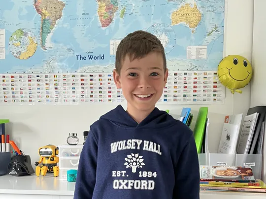 Happy student wearing a Wolsey Hall Oxford hoodie, standing in front of a colourful world map at home