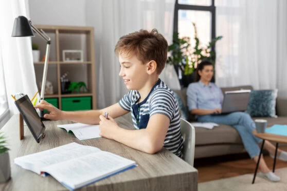 A homeschooling young boy with short brown hair sitting at a desk and studying on a tablet device. His mother is sitting on a sofa in the background.
