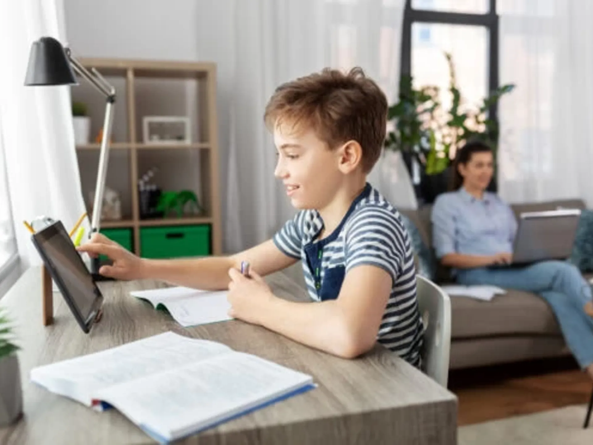 A homeschooling young boy with short brown hair sitting at a desk and studying on a tablet device. His mother is sitting on a sofa in the background.