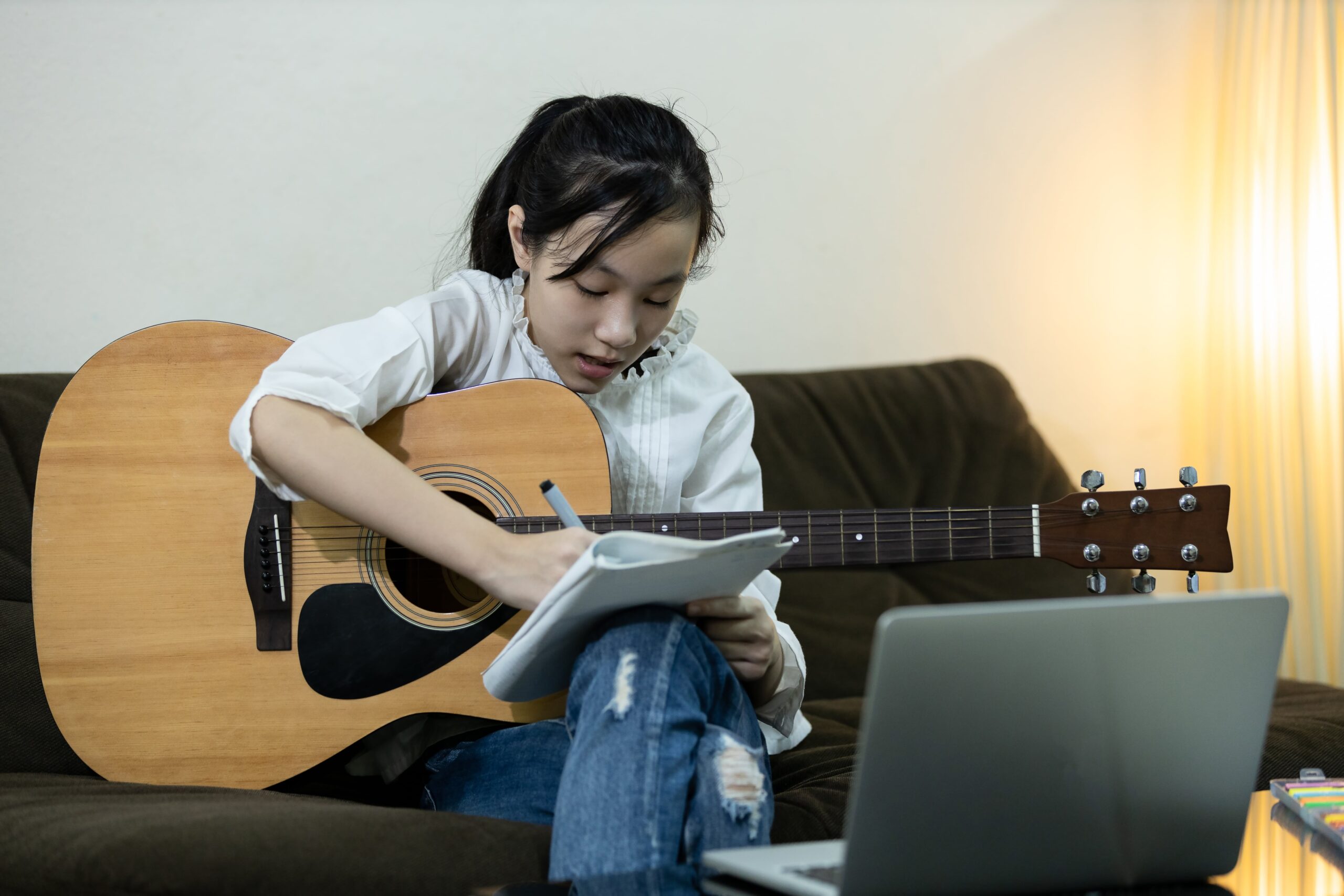 Girl writing music notes in a notebook while holding an acoustic guitar during an online lesson.