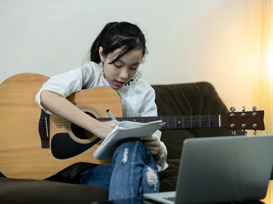 Girl writing music notes in a notebook while holding an acoustic guitar during an online lesson.