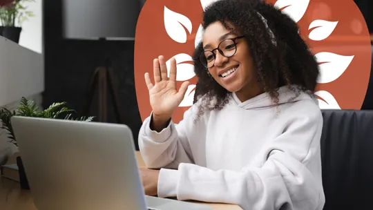 A young person with glasses and headphones, smiling and waving at a laptop screen, engaged in an online video call.