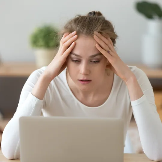 A stressed and anxious secondary student holds her head in her hands while looking at her laptop screen, representing anxiety.