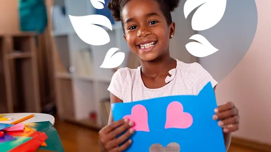 A homeschooling young girl smiling holding up a creative craft picture with pink hearts on blue card.