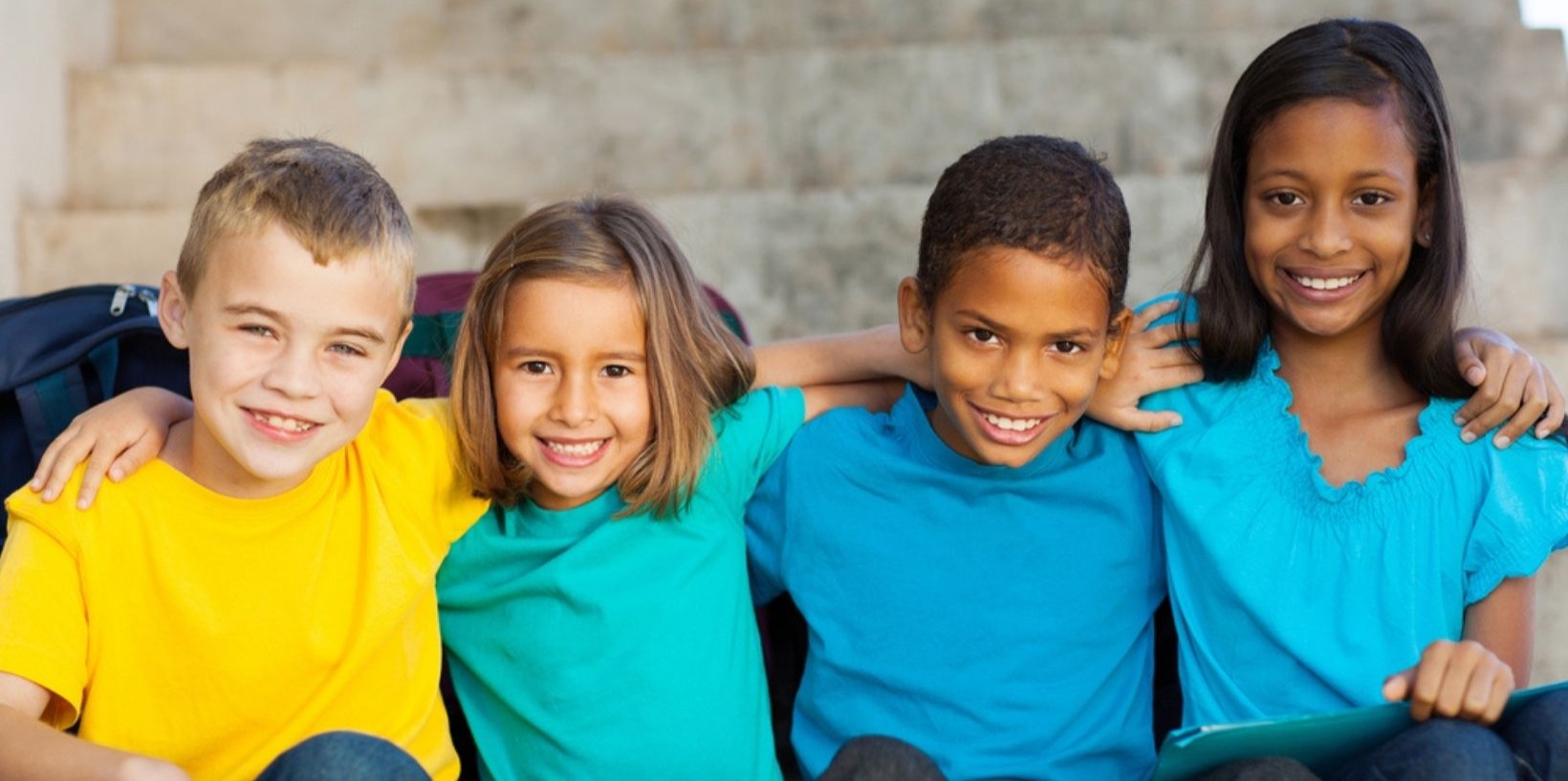 Four children sitting outdoors with arms around each other, smiling and enjoying friendship