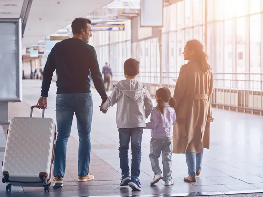 Family of four walking hand in hand through airport with travel bags