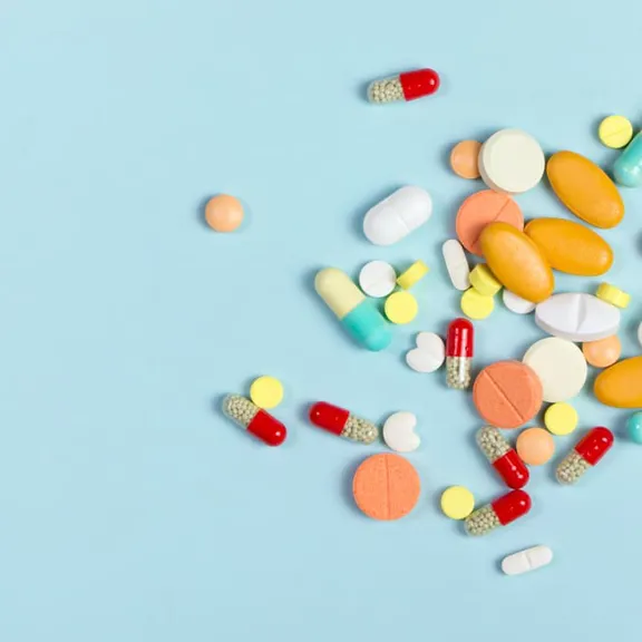 A flat lay of various-sized pills and capsules in different colors, including orange, yellow, red, white, and blue-green, scattered on a solid light blue background.