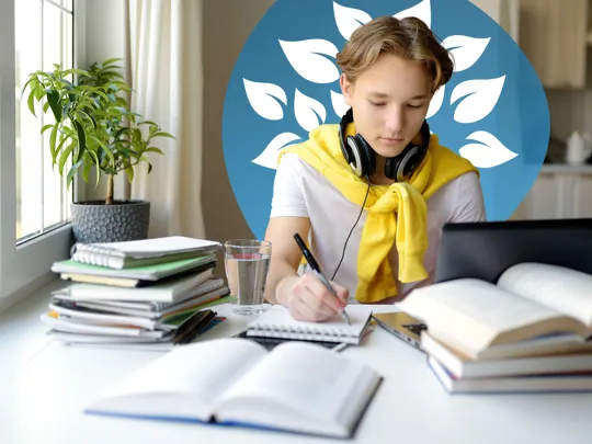 A teenage student with headphones sits at a desk at home, writing in a notebook while studying online with a laptop and books.