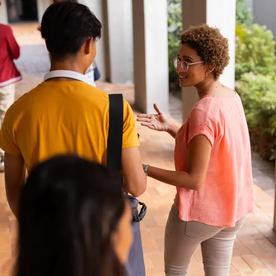 A friendly female teacher or tutor wearing glasses and a pink shirt is walking and talking animatedly with a male student in a yellow shirt in a university or school hallway lined with brick-colored tiles and white pillars. Other students are walking in the background.