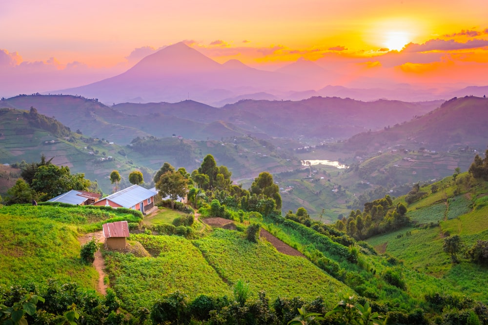 A vibrant sunset over rolling green hills and valleys in Uganda, with small buildings and mountains in the distance.