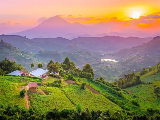 A vibrant sunset over rolling green hills and valleys in Uganda, with small buildings and mountains in the distance.