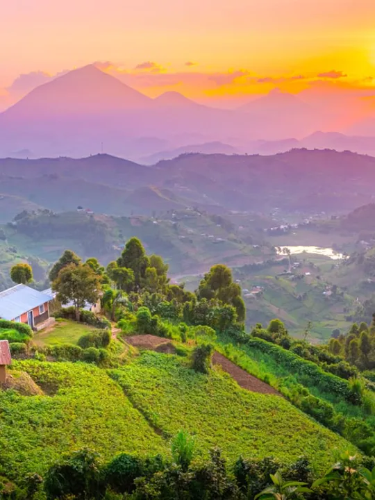 A vibrant sunset over rolling green hills and valleys in Uganda, with small buildings and mountains in the distance.