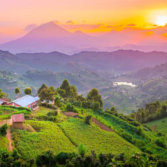 A vibrant sunset over rolling green hills and valleys in Uganda, with small buildings and mountains in the distance.