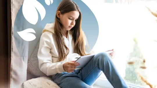 Young girl with long hair reading on a tablet while sitting on a cushion by a bright window.