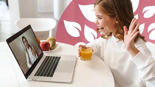 Smiling girl waving at a laptop screen during a video call, with a glass of juice in hand.