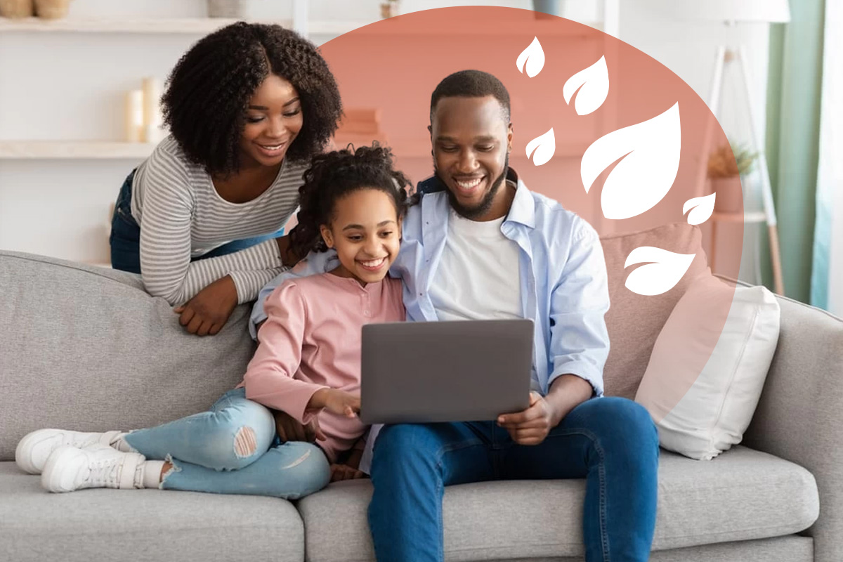A homeschooling mum, dad and young daughter in their living room smiling at a laptop. The dad and daughter are sat on the sofa, whilst the mother is leaning on the back of it.