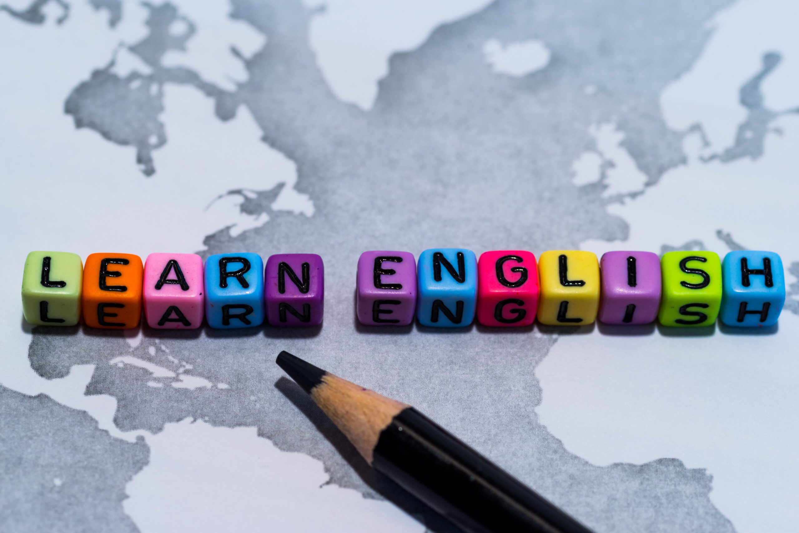 Colourful letter beads spelling ‘Learn English’ on a monochrome map with a pencil in the foreground.