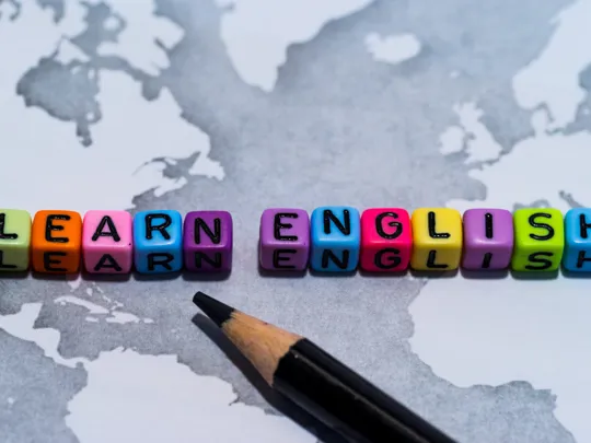 Colourful letter beads spelling ‘Learn English’ on a monochrome map with a pencil in the foreground.