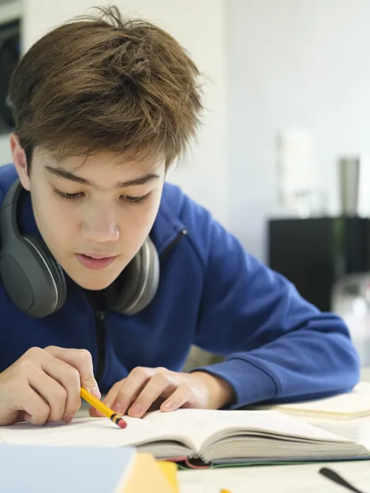 Young student with headphones studying a textbook and taking notes at a desk while learning online at home. This image represents inclusive, flexible digital education for students with SEND (Special Educational Needs and Disabilities) through Pamoja Education.