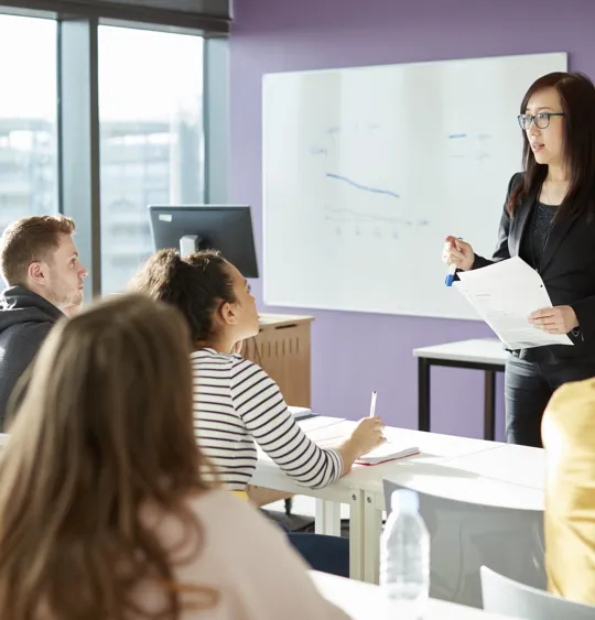 Pamoja teacher holding papers addressing learners in a modern classroom.