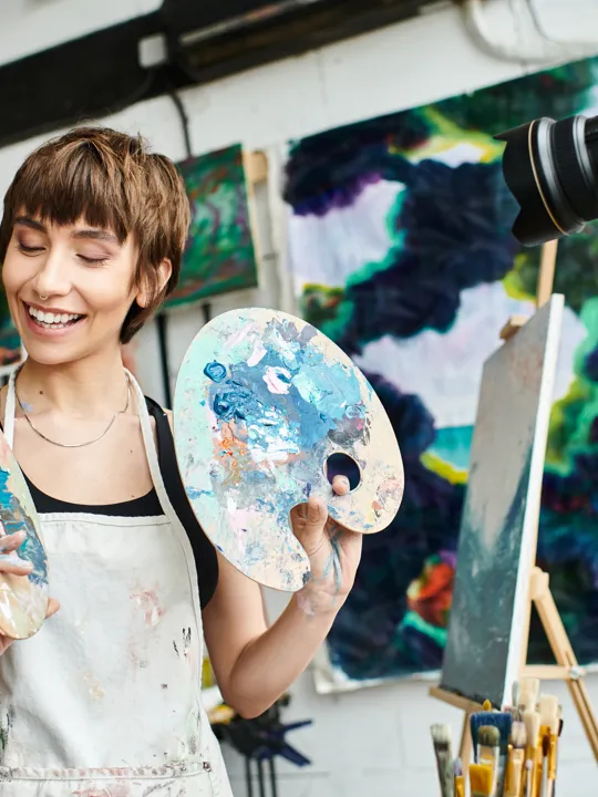 A smiling young woman in an art studio, holding two paint palettes, with a camera on a tripod, easel, and abstract paintings in the background.