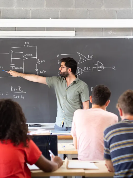 a male teacher explaining a circuit diagram on a chalkboard to a class of high school or university students.