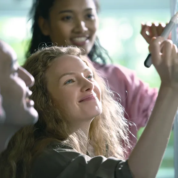 Four smiling colleagues collaborating on an idea; a woman with long blonde hair writes on a clear whiteboard while others look on.