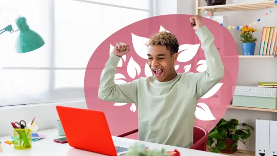 Teen boy celebrating success in front of a red laptop at a tidy home study desk.