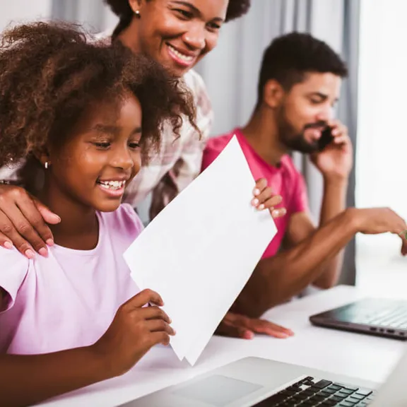 Young child smiling at laptop whilst holding a piece of paper.