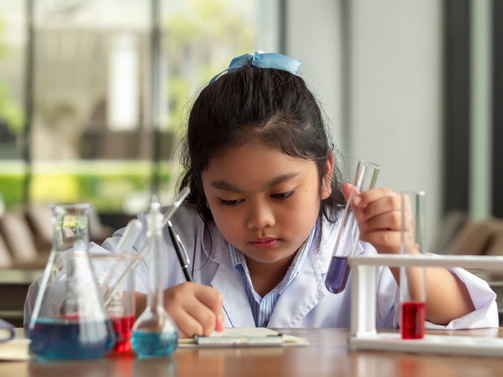 Focused student in lab coat writing notes while holding a test tube, surrounded by colourful beakers