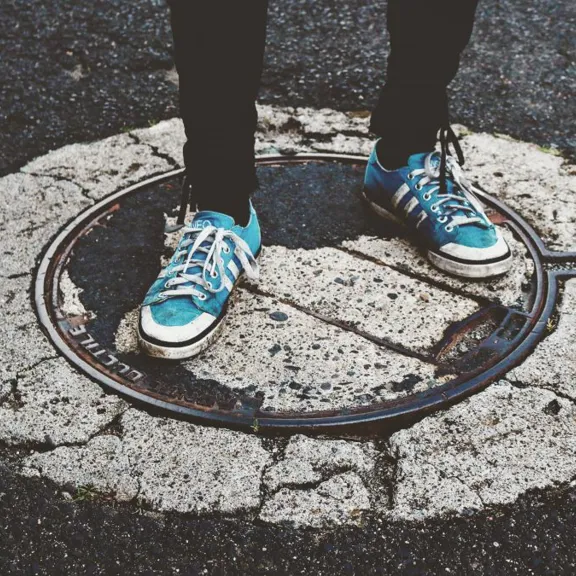 A low-angle shot showing a person's legs and feet standing on a manhole cover in the middle of a street. The person is wearing black trousers and a pair of blue sneakers with white laces. The manhole cover is surrounded by cracked concrete and asphalt.