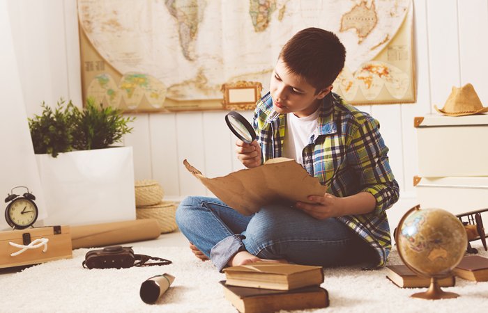 Young boy examining a historical map with a magnifying glass, surrounded by books and a globe.