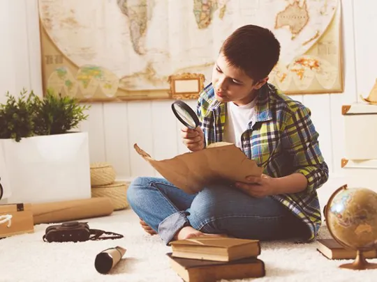 Young boy examining a historical map with a magnifying glass, surrounded by books and a globe.