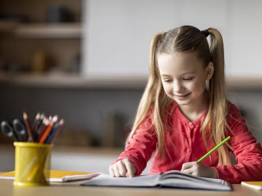 Smiling primary-age girl with pigtails happily writing in a notebook, representing engaging homeschooling for primary children