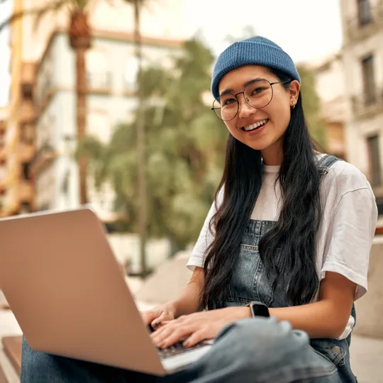A young woman in a blue beanie and overalls smiles while working on a rose gold laptop outdoors on a sunny city street for online learning