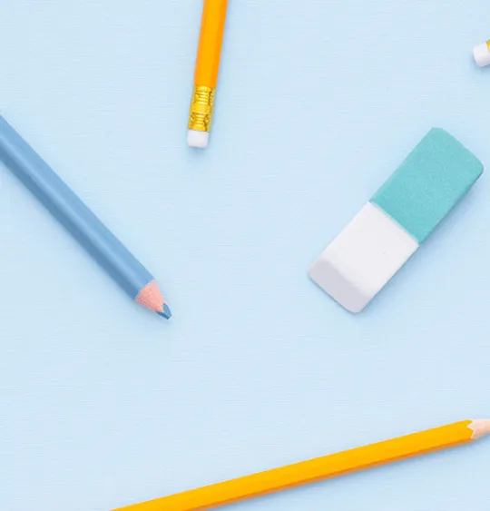 A flat-lay image of various school supplies scattered on a light blue background. The supplies include yellow and colored pencils, a blue eraser, a ruler, and washi tape.