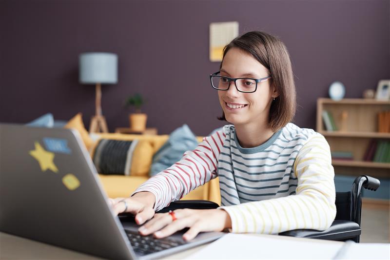 A smiling teenage student in a wheelchair engages happily with her online studies from home.