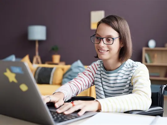 A smiling teenage student in a wheelchair engages happily with her online studies from home.