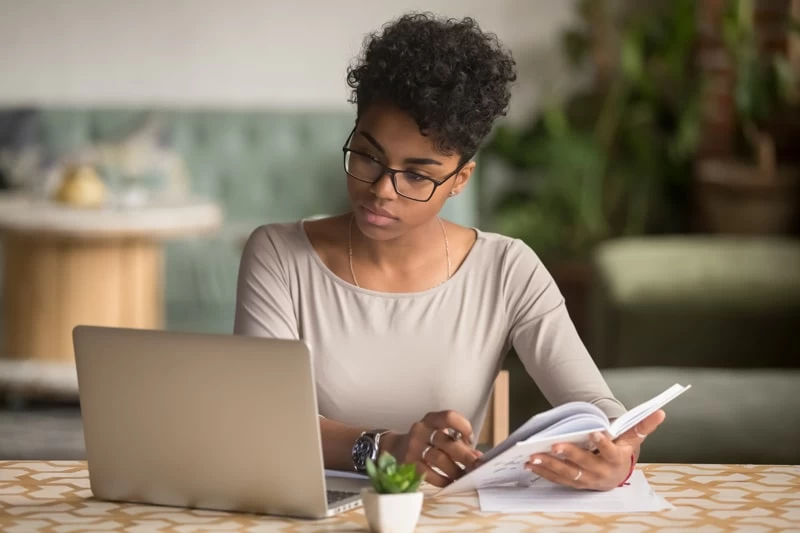 A woman studying on a laptop with a notepad in hand. 