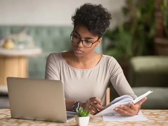 A woman studying on a laptop with a notepad in hand.