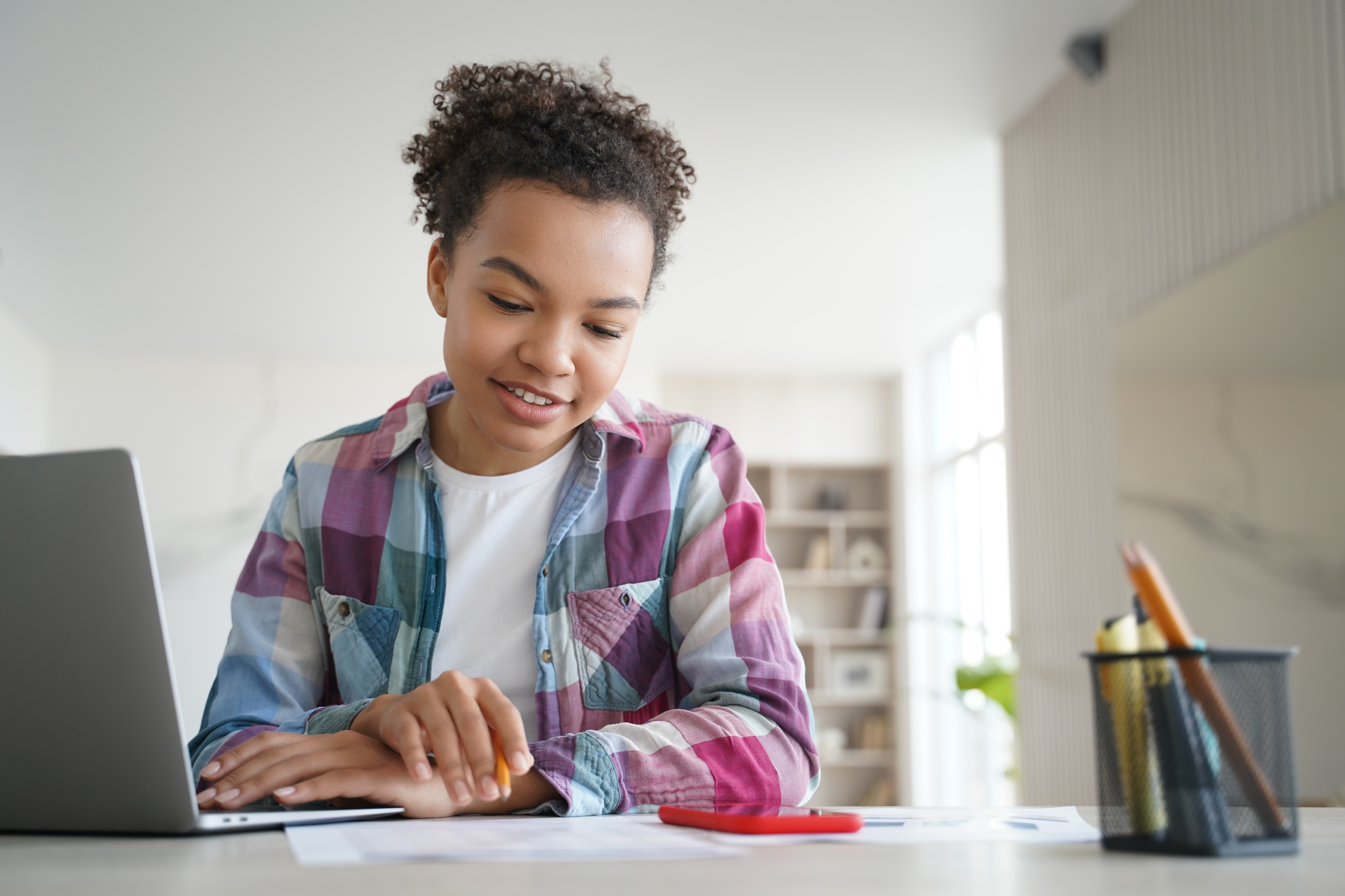 A young student in a plaid shirt sits at a desk, focused on their laptop screen while holding a pencil, engaged in online learning in a bright room.