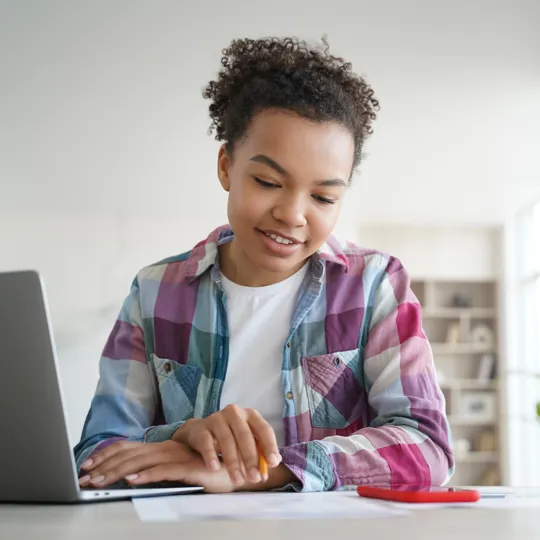 A young student in a plaid shirt sits at a desk, focused on their laptop screen while holding a pencil, engaged in online learning in a bright room.