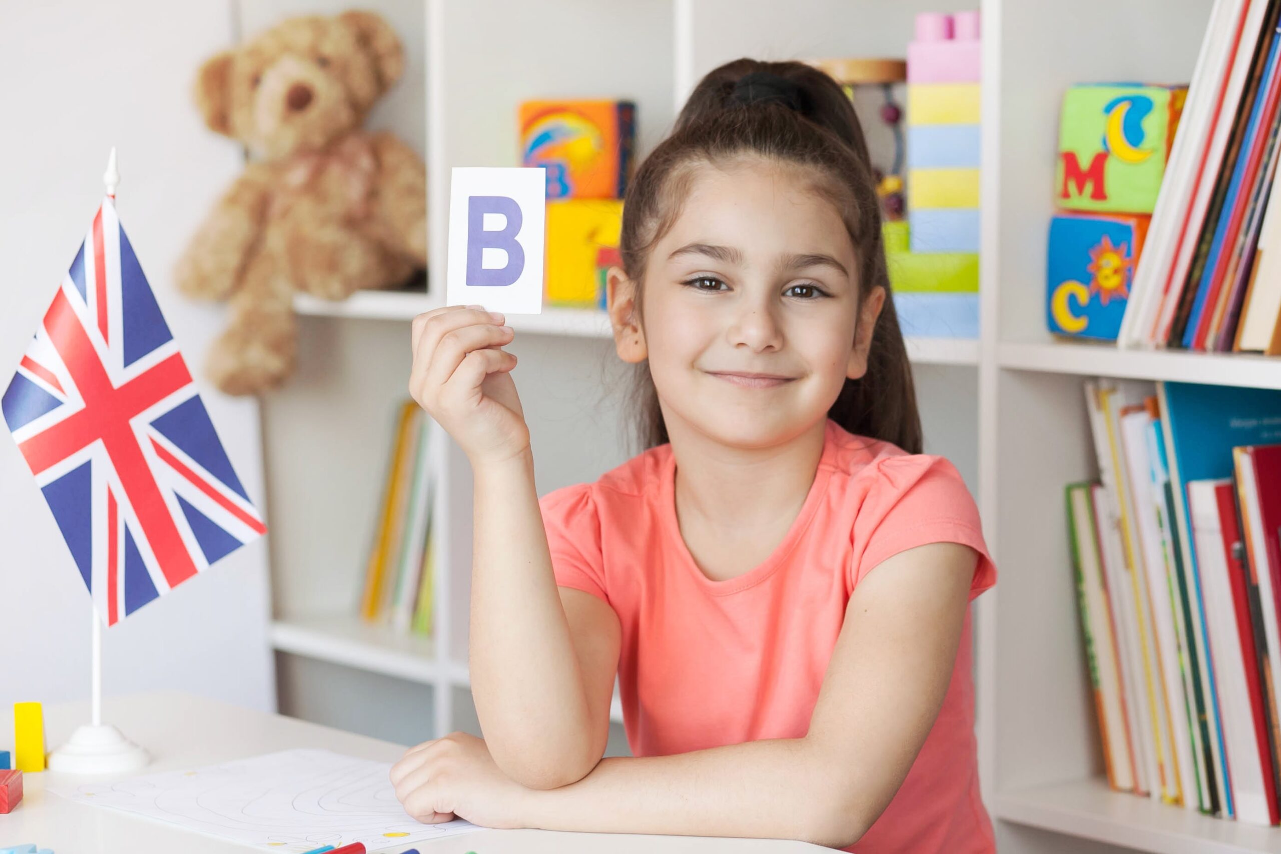 Smiling girl in a classroom holding up a flashcard with the letter B beside a Union Jack flag.
