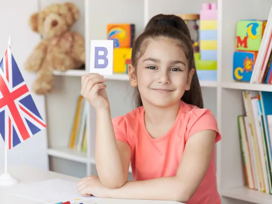Smiling girl in a classroom holding up a flashcard with the letter B beside a Union Jack flag.