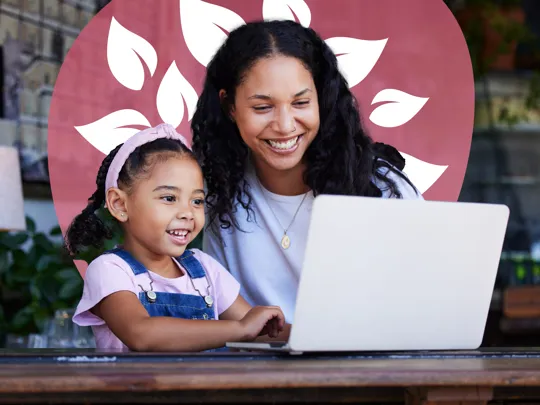 An adult and a young girl smiling and looking at a laptop together, appearing to be in a cafe setting.