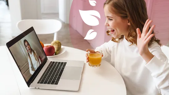 Smiling girl waving at a laptop screen during a video call, with a glass of juice in hand.