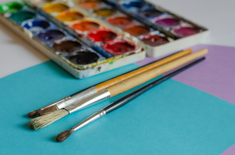 Watercolour paint palette and three paintbrushes resting on coloured paper, ready for a creative art activity