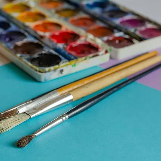 Watercolour paint palette and three paintbrushes resting on coloured paper, ready for a creative art activity